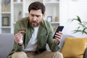 Confused man sitting on a couch holding a phone and a credit card.